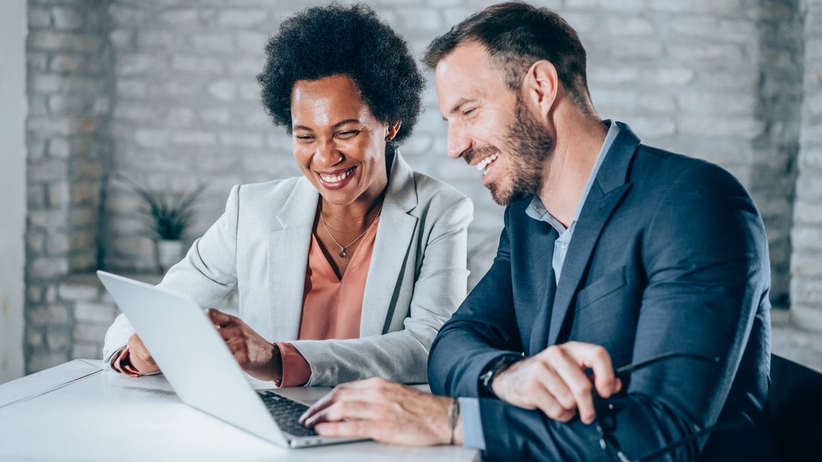 Shot of a two confident business persons sitting on a desk in the office. Businessman and businesswoman in meeting using laptop and discussing business strategy. Business coworkers working together in the office.